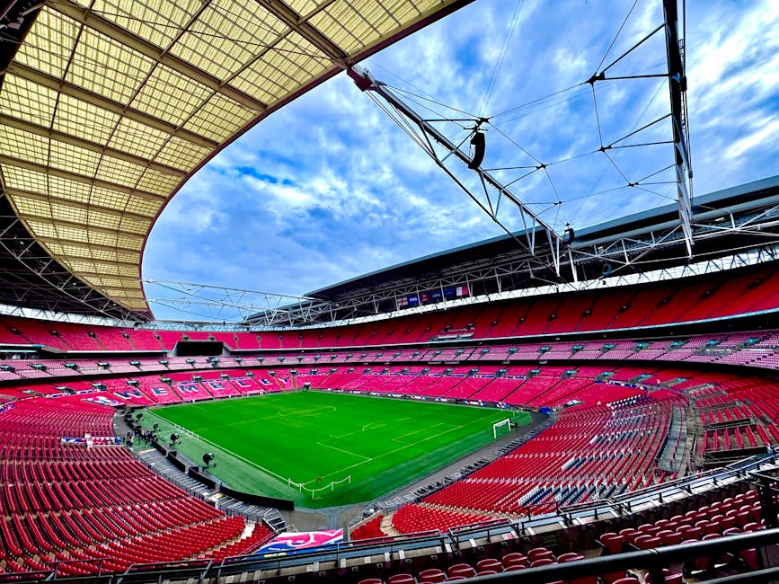 Interior view of a large, covered football stadium with a green grass pitch and seating sections in shades of red. The protective roof structure extends over the stands, with steel support beams and overhead electrical wiring visible. Cardboard boxes, wrapped furniture, and packing materials are arranged along one side of the stadium, suggesting preparations for a home relocation or furniture transport. Some boxes and wrapped items are positioned on the pavement near an open doorway leading into the interior, while staff members are seen lifting and carrying various items onto or off a vehicle outside the building, with a delivery truck or removal van partially visible in the background. The scene captures the process of loading household goods during a packing and moving operation, consistent with professional removal services such as those provided by Twickenham Movers, in a well-lit environment under partly cloudy skies.