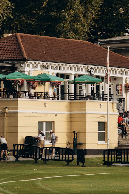 A street-level view of a two-storey building with a red-tiled roof and cream-colored exterior walls, situated near Twickenham Stadium. The upper terrace extends into an outdoor seating area with green umbrellas featuring the Heineken logo, supported by black metal railings and accessible via stairs on the right side. Several people are gathered on the terrace, some seated at tables, while others are walking or standing near the entrance of the building below. The lower level has small windows, and outside on the lawn, there are black benches, a trash bin, and a small group of individuals. Tall trees with dense foliage shade the scene, and a flagpole with a white flag is visible near the building. The overall setting depicts a lively atmosphere in the area associated with outdoor dining, possibly during an event or a busy day near Twickenham Stadium, with Twickenham Movers involved in the professional packing and home relocation services related to nearby moving projects.